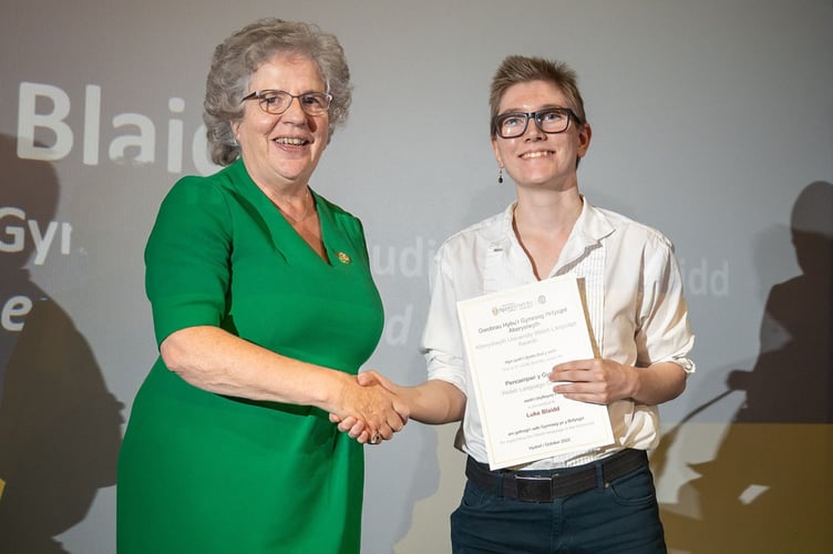 Y myfyriwr is-raddedig Luke Blaidd yn derbyn gwobr Pencampwr y Gymraeg oddi wrth yr Athro Elizabeth Treasure, Is-Ganghellor y brifysgol. Ac yntau wedi ei eni yn Stockport a’i fagu yn Manceinion, Luke yw’r cyntaf o’i deulu i siarad Cymraeg.