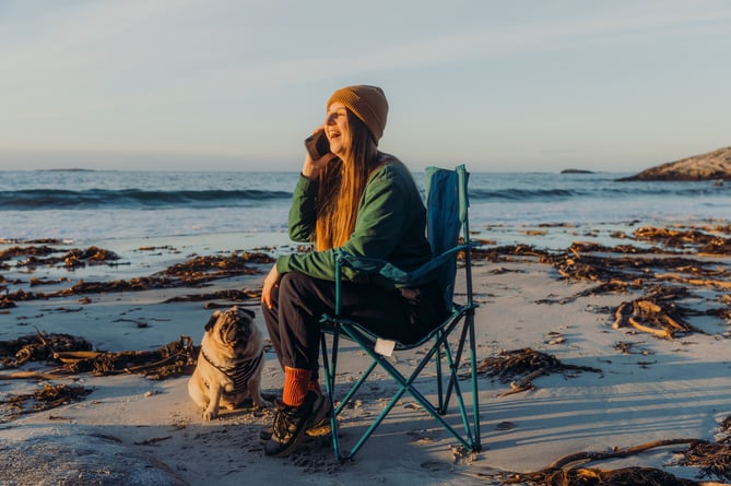 Happy woman in yellow hat with her cute pug enjoying vacation by the seashore of Norway and doing business by phone during the freelance work