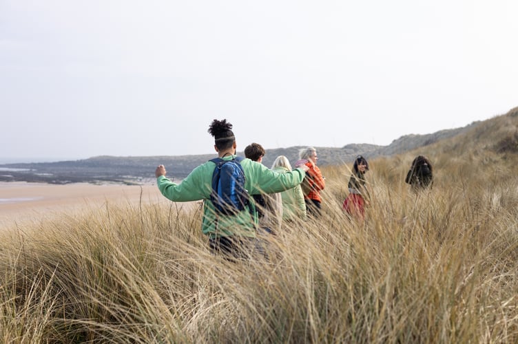 Wide rear view of a medium group of young and mid-adult friends dressed in warm, casual outdoor clothing enjoying a day out in Newton-by-the-Sea, Alnwick. They have their arms raised wide as they walk through the scenic British countryside, surrounded by rolling dunes and natural landscapes. 

Videos are available similar to this scenario.